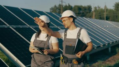 İki teknisyen, bir kadın ve bir adam, güneş panelleri arasında yürürler ve bir güneş istasyonunun işleyişini tartışırlar. Güneş enerjisi santralinde yeşil enerji kavramı. Yüksek kalite 4K görüntü.