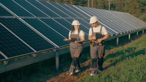2人の若い技術者 1人の女性と1人の男が太陽光発電パネルの列を歩き 太陽と緑のエネルギーの仕事を一緒に話し合った 作業効率について 高品質の4K映像 — ストック動画