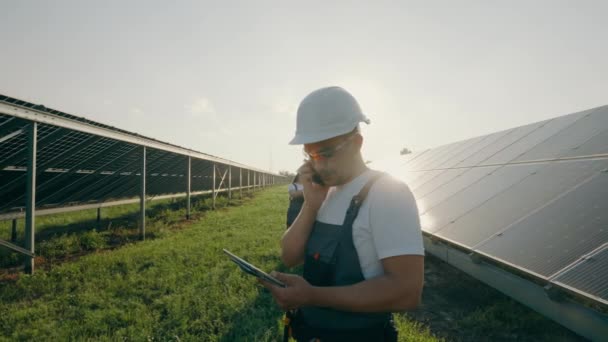 太陽光発電所に立っている間 携帯電話で話している監督エンジニアの肖像画 バックグラウンドでソーラーパネルを検査する労働者 高品質の4K映像 — ストック動画