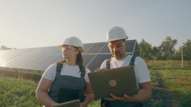 Sert şapkalı ve dizüstü bilgisayarlı bir erkek ve kadın dışarıdaki güneş panellerinin arka planına karşı çalışma görevlerini tartışıyorlar. Yatırımcıyla konuşan kadın çevre mühendisi. Yeşil elektrik kavramı.