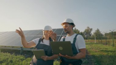Güneş panellerini kontrol ederken güvenlik kaskları ve yelekleriyle konuşan ve el kol hareketi yapan becerikli mühendislerin arka görüşü. Bir erkek ve kadın ortak bir yeşil enerji projesi olan 4K üzerinde işbirliği yapıyorlar.