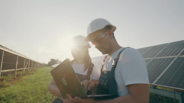 一位头戴硬帽 手持笔记本电脑的男男女女正在户外太阳能电池板的背景下讨论工作任务 女环境工程师与投资人交谈 — 图库视频影像
