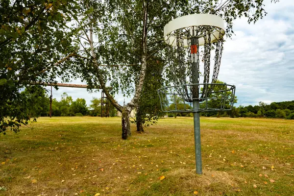 Bir disk golf sepeti huş ağaçları arasındaki açık bir çayırda duruyor. Arka planda, endüstriyel bir bitkinin eski kalıntıları geçmişe işaret ediyor, ve sahne sakin ve sportif görünüyor..