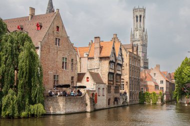 A line of structures near a river with a distant church, blending into the natural landscape surrounded by water, trees, and clouds in the sky