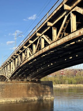 Paslı çelik köprü huzur nehri boyunca uzanır, parlak mavi gökyüzünün altındaki sudaki eşsiz mimari özellikleri ve yansımaları vurgular.