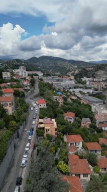 Bulutlu bir gökyüzünün altında yeşil ağaçlar ve tepelerle çevrili kırmızı kiremitli çatıları olan yerleşim alanının havadan görüntüsü. Sahne, kentsel ve doğal unsurları uyum içinde yakalıyor. Web sitesi arkaplan videosu