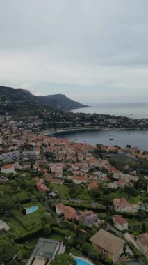 Renkli evleri, yeşil tepeleri ve sakin bir denizi olan kıyı kasabasının havadan görünüşü. Gökyüzü bulutlu, sakin bir atmosfer yaratıyor..