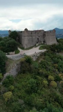 Yeşilliklerle çevrili antik bir taş şatonun havadan görünüşü. Kalede, girişe giden dolambaçlı bir yolu olan yüksek duvarlar ve kuleler yer alıyor..