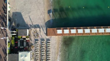 Riviera 'nın gök mavisi suları ve Fransa' nın Cannes şehrinin havadan görünüşü. Sahnede kumlu bir sahil, güneş pansiyonları ve denize uzanan ahşap bir iskele yer alıyor. Web sitesi geçmişi, turizm 