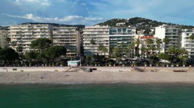 Fransa, Cannes 'ın gök mavisi kıyı şeridi ve şehir manzarası. Sahne boyunca kumlu plajlar, palmiye ağaçları ve modern binalar yer alıyor. Kıyı yaşam tarzları, tatil reklamları