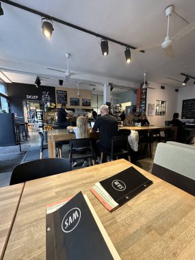 Varşova, Polonya - 22 Mayıs 2025: Ahşap masalı rahat kafe sahnesi, içeceklerin tadını çıkaran müşteriler, ve SAM menüsü görünür, sosyalleşmek için sıcak bir ortam yaratıyor