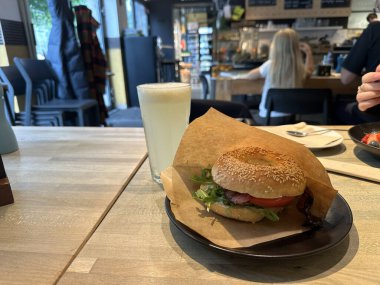 Varşova, Polonya - 22 Mayıs 2025: Marul ve domatesli lezzetli susamlı burger, sıcak bir kafe atmosferinde soğuk bir içecekle eşleştirildi