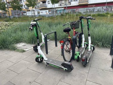 Varşova, Polonya - 22 Mayıs 2025: Elektrikli scooterlar ve bir bisiklet yemyeşil çimenler ve çağdaş mimariyle canlı bir şehir ortamında park halindedir.