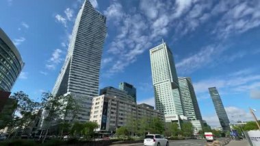 Gökdelenler açık mavi gökyüzüne karşı, ağaçlar ve araçların canlı şehir atmosferine eklediği modern mimari tarzları ve şehir manzarasını vurguluyor.