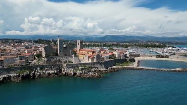 Deniz kenarında antik taş kuleleri olan, tepeler ve dinamik bulut oluşumlarıyla çerçevelenmiş bir marina ve kayalık kıyı şeridini gösteren bir kasabanın sahili.
