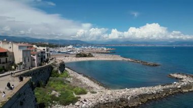 Kıyı manzarasında kumlu bir sahil, berrak sular ve uzaktaki yatlar, dağlarla çevrili ve hareketli bulutlar, sakin bir yaz havası uyandırıyor.