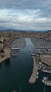 Canlı bir marina sahnesinde, manzaralı mimari ve dalgalı tepeler tarafından çerçevelenen ve canlı sahil atmosferini yansıtan çok sayıda tekne ve yat sergileniyor.