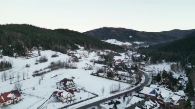 Manzaralı hava perspektifi karla kaplı evleri ve kıvrımlı yolları, yemyeşil ağaçların arasında sergiliyor, kırsal bir kışın sükunetini vurguluyor.