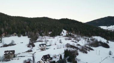 Hava perspektifi, karla kaplı tepeler, dağınık evler ve ebedi yeşil ağaçlarla dolu bir kış manzarasını sergiliyor.