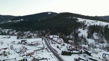 Manzaralı hava perspektifi karla kaplı evleri ve kıvrımlı yolları sergiliyor yemyeşil ağaçların arasında, kırsal alanda kışın huzurlu özünü vurguluyor.