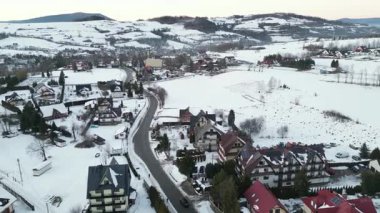 Karla kaplı tarlaları, büyüleyici evleri ve dolambaçlı yolları olan, kışın kırsal bir arazinin sükunetini yakalayan bir kış manzarasının çarpıcı hava perspektifi.