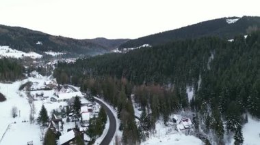 Karlı kış manzarası yukarıdan çekildi, büyüleyici evler, dolambaçlı yollar ve sık yeşil ormanlar sergileniyor, sakin ve resimli bir manzara yaratıyor.