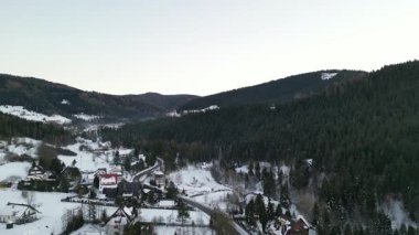 Kış manzaraları kar ve yemyeşil ağaçlarla çevrili büyüleyici bir köy, arka planda tepeler, sükuneti ve doğal güzelliği çağrıştırıyor.