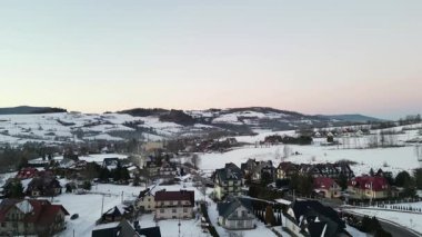 Karla kaplı çatılar, yuvarlanan tepeler ve uzak dağların yer aldığı pitoresk bir kış köyü sahnesi manzarada huzurlu ve sakin bir ortam yaratıyor.