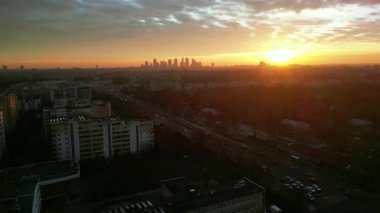Gün batımında şehir silueti, yollar ve binalar, sıcak renklerle gökyüzünü boyuyor, sakin ve güzel bir şehir manzarası yaratıyor.
