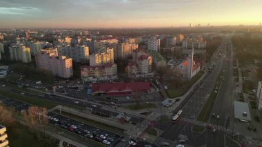 Gün batımında yukarıdan çekilen şehir manzarası konut binalarını, işlek caddeleri ve araçları vurguluyor, canlı bir akşam atmosferi yaratıyor.