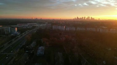 Gün batımında şehir manzarası, renkli bir gökyüzü ve uzak bir ufuk çizgisi ile yerleşim binalarını ve yolu aydınlatıyor, sakin bir şehir atmosferini çağrıştırıyor.