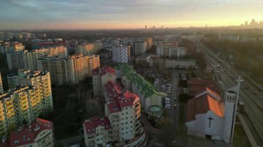 Gün batımında yukarıdan çekilen kentsel manzara, konut binaları, yollar ve canlı şehir atmosferini gösteren uzak bir ufuk çizgisi içeriyor.