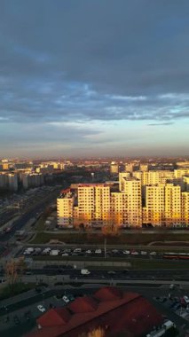 Gün batımında kentsel alanın manzarası, konut binalarının ışıklandırılması, yoğun yollar ve renkli bir gökyüzü, barışçıl ama dinamik bir şehir havasını çağrıştırıyor.