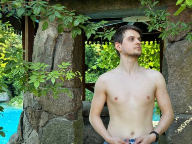 Genç adam canlı yeşil yapraklarla çevrili taştan bir yapının yanında poz veriyor. Arka planda sakin bir havuz var. Bu da dışarıda huzurlu bir atmosfer yaratıyor.