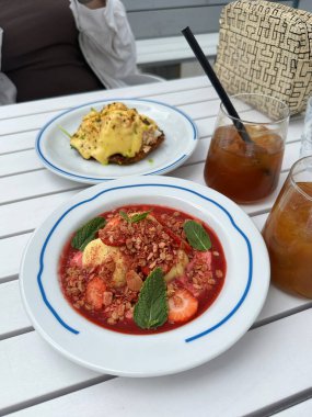 Çilekler, muzlar ve nane yapraklarıyla süslenmiş granola içeren canlı bir tatlı. Lezzetli bir tabağın yanında beyaz bir tabağa konmuş ve serinletici buzlu içecekler.