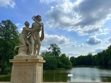 Sersemletici mermer heykel, doğadaki sevgi ve uyumu sembolize eden canlı yeşillik ve dramatik gökyüzü ile sakin bir gölün yanında kucaklaşan iki figürü tasvir ediyor.