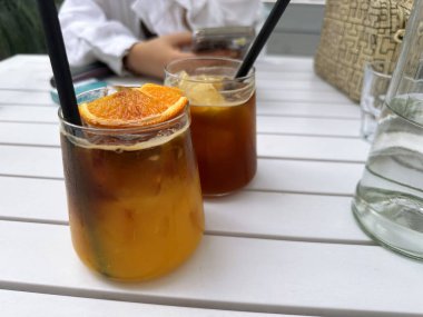 Buzlu içecekler, turuncu süslemeler ve pipetlerle dolu cam kavanozlarda, beyaz ahşap bir masaya yerleştirilip, sosyal etkinlikler için canlı ve davetkar bir ortam yaratıyor.