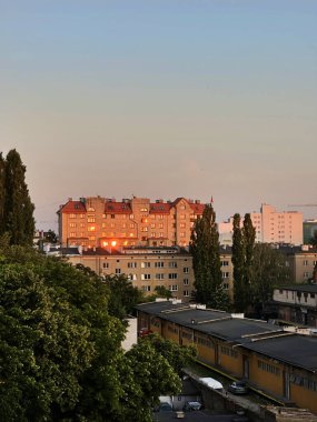 Günbatımında şehir manzarasında aydınlık pencereli binalar, ön planda yeşillik ve kentsel yaşamın özünü yansıtan sakin bir gökyüzü yer alıyor.
