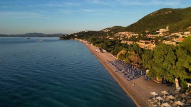 Corfu Island, Greece. Coastal scene captures the gradual shift from calm waters to a sandy beach, featuring colorful umbrellas and lush trees, creating a peaceful atmosphere