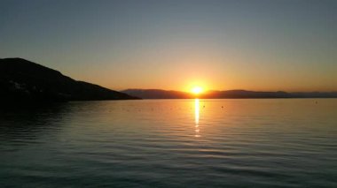 Tranquil sunrise scene unfolds over still waters, with golden light reflecting on the surface and distant hills creating a serene coastal atmosphere