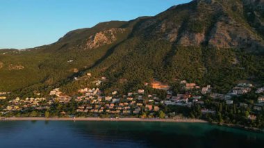 Aerial perspective reveals a tranquil coastal scene with a vibrant beach, lush hills, and a charming village, highlighting the serene evening ambiance and natural beauty