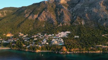 Aerial perspective reveals a coastal village embraced by mountains, with clear waters reflecting the landscape, highlighting the peaceful coexistence of nature and architecture