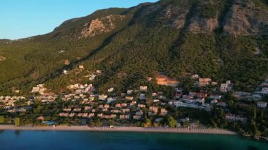 Aerial perspective reveals a coastal village surrounded by mountains and clear waters, highlighting charming homes and a peaceful beach, embodying serene island living
