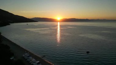 Sakin suların üzerinde sakin bir gün batımı beliriyor, sahneyi aydınlatan altın yansımalarla, yumuşak dalgalar kıyıya vururken, sakin sahil atmosferini güçlendiriyor.