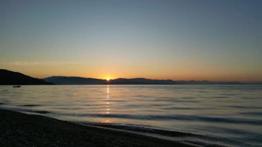 Durgun suların üzerinde büyüleyici bir gün doğumu uzak tepelere sıcak bir ışık tutuyor ve sakin bir atmosfer yaratarak sakin bir sahil manzarasının güzelliğini gözler önüne seriyor.