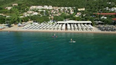 Paddleboardcular, güneşli şezlonglar ve güneşliklerle çevrili bir plaj tarafından çerçevelenmiş sakin sularda süzülerek rahatlatıcı ve maceralı bir atmosfer yaratırlar.