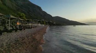 Güneşli sahil şeridi, şezlonglar ve yumuşak dalgalar ile sakin sahil manzarası gelişiyor, rahatlamayı davet ediyor ve kıyı yaşamının huzurlu atmosferini gösteriyor.