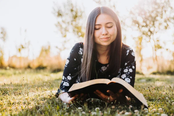 Young believing girl lying on the grass and studying her bible, in the field at sunset. Copy space. High quality photo