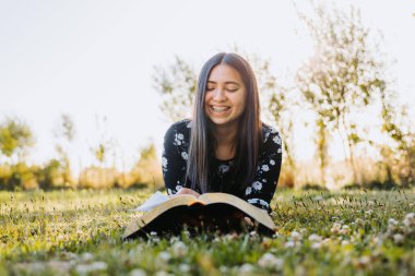 Young smiling woman, believer, studying her bible while lying on the grass, in the field at sunset. High quality photo
