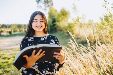 Smiling young religious girl reading her bible, outside in the field at sunset. Spiritual revival.. High quality photo